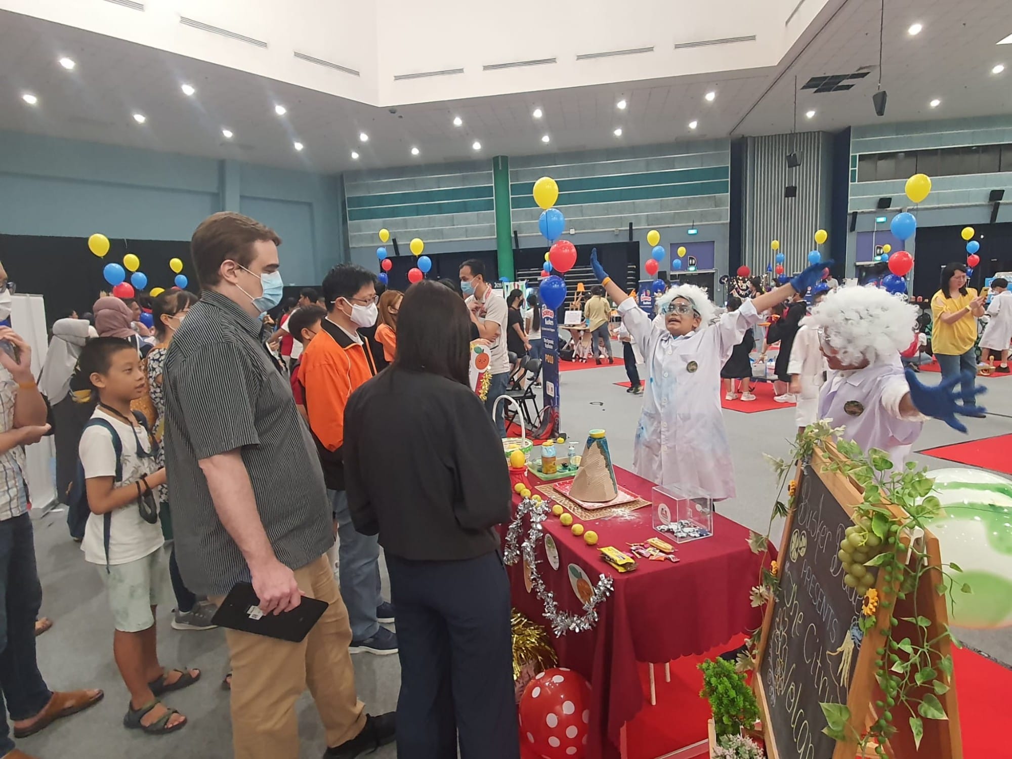 Children in lab coats and white wigs present a science fair project to visitors in a large hall with colorful balloons.